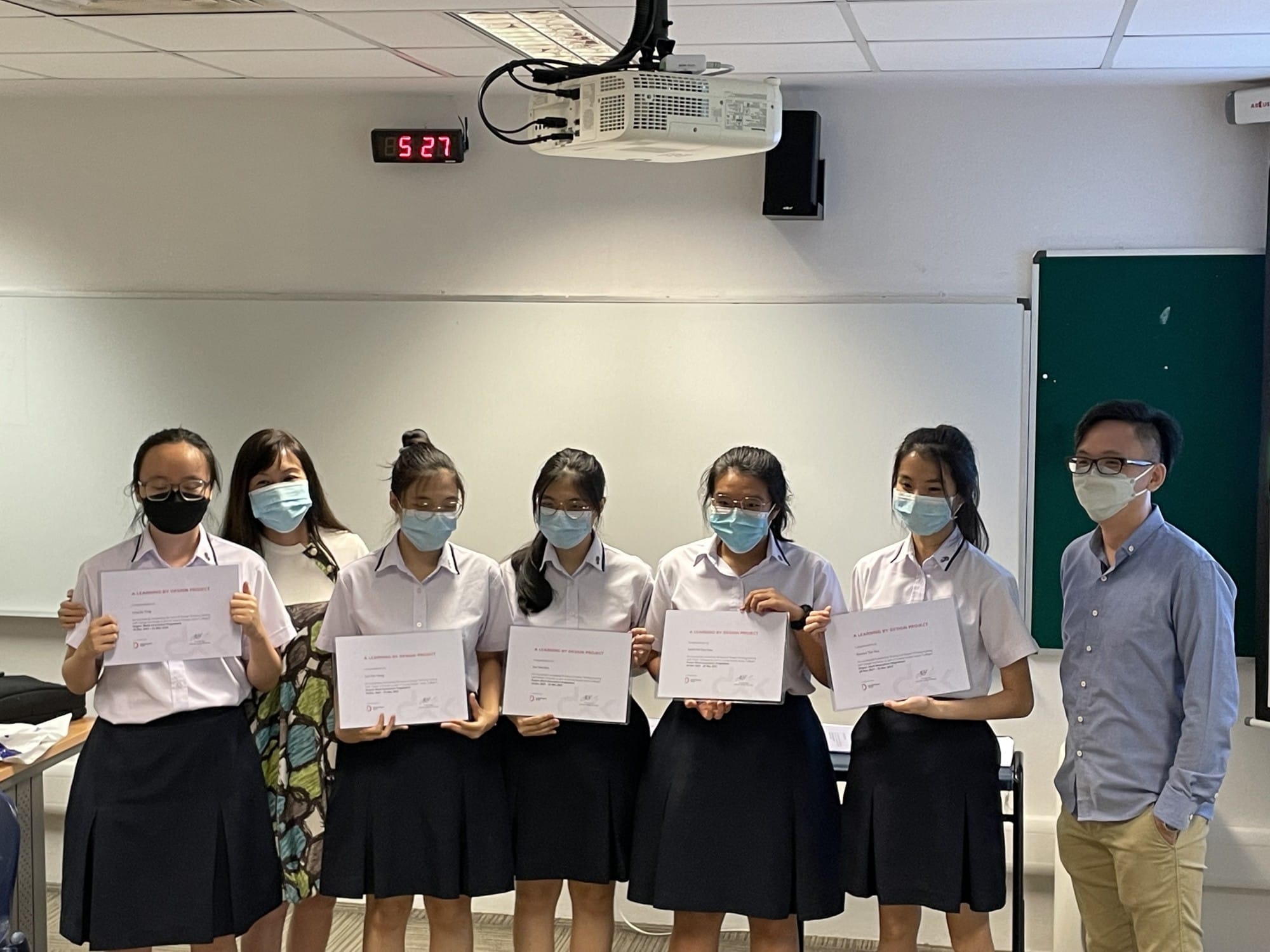 Seven masked people stand in a classroom, with five students in uniform holding certificates alongside two adults.